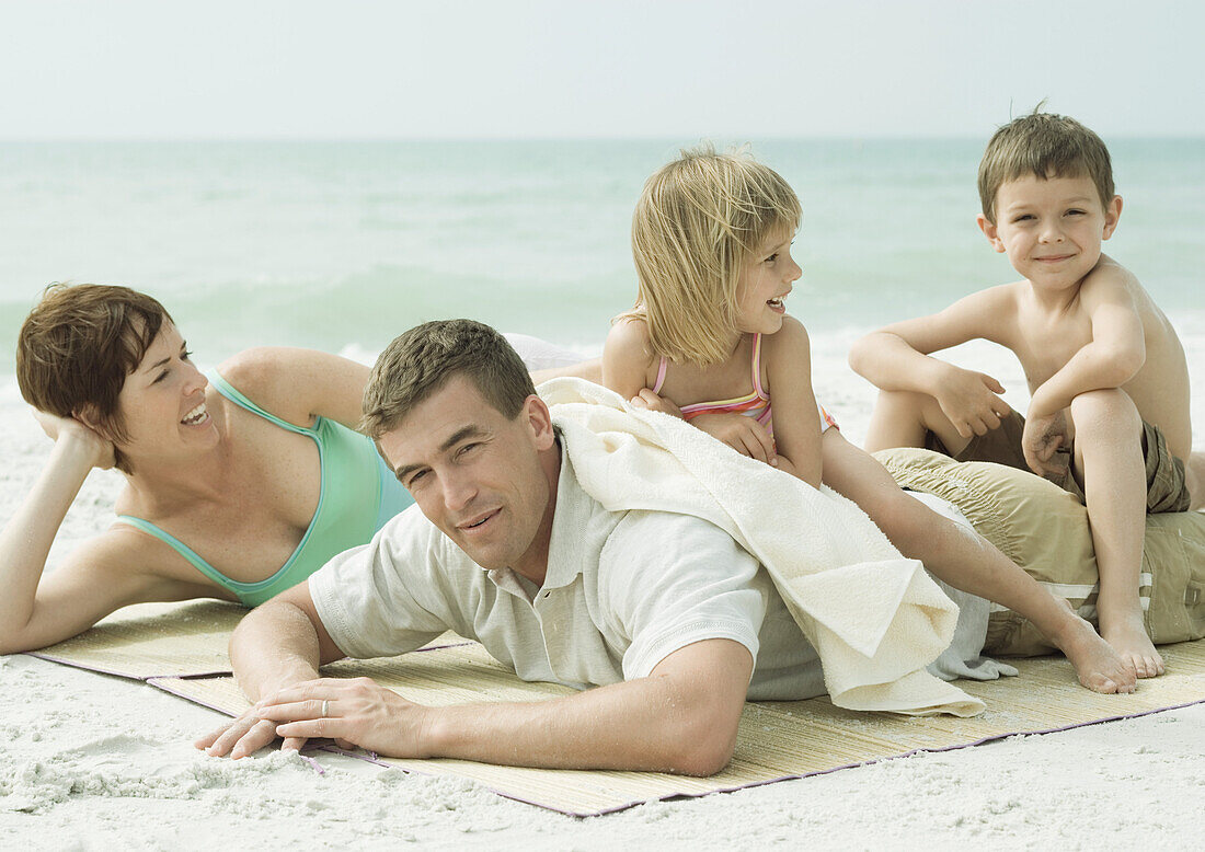 Family at the beach, parents lying on mat while children sit on father's back