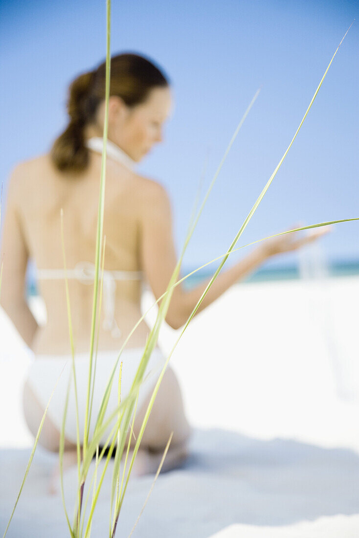 Young woman sitting on beach, focus on dune grass in foreground