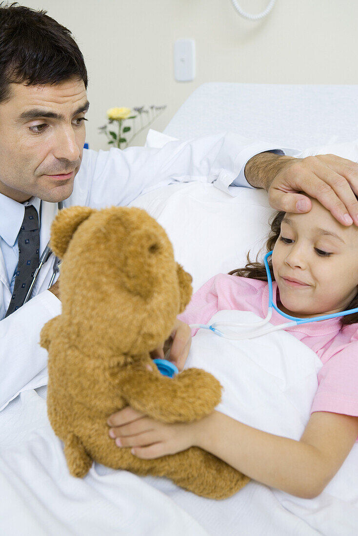 Child holding toy stethoscope to teddy bear, doctor feeling child's forehead
