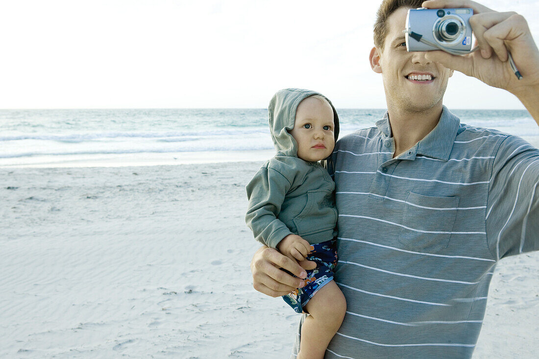Man holding baby and using camera on beach