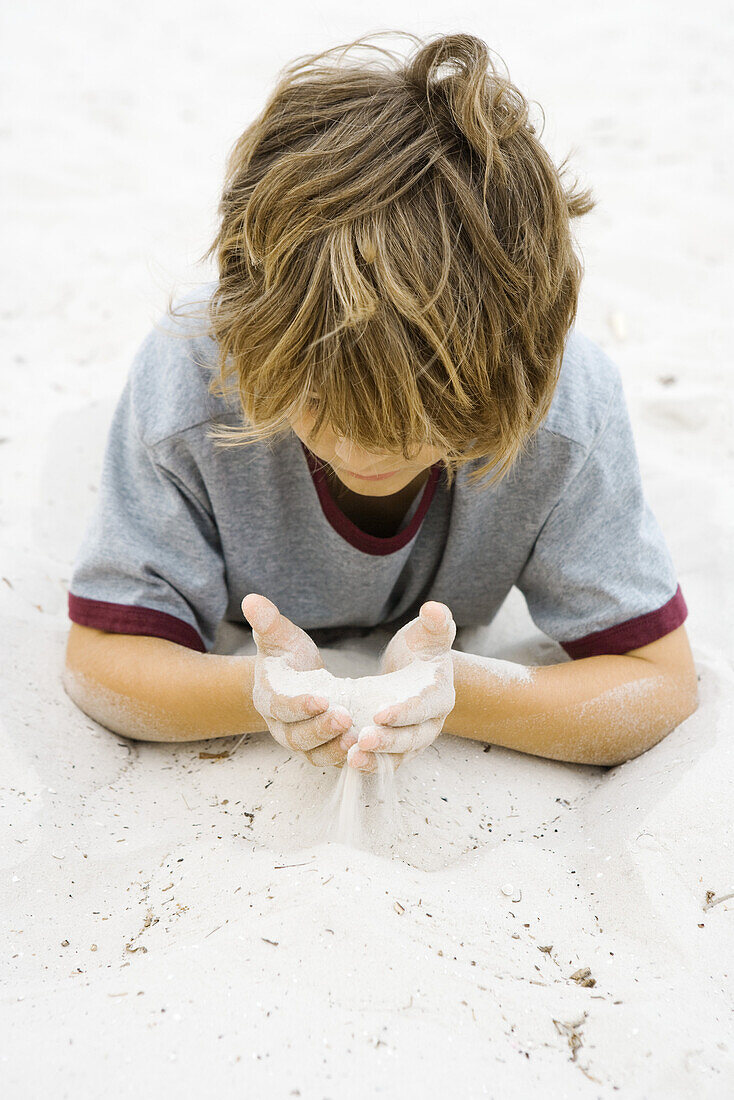 Boy lying on the ground, looking down at handful of sand