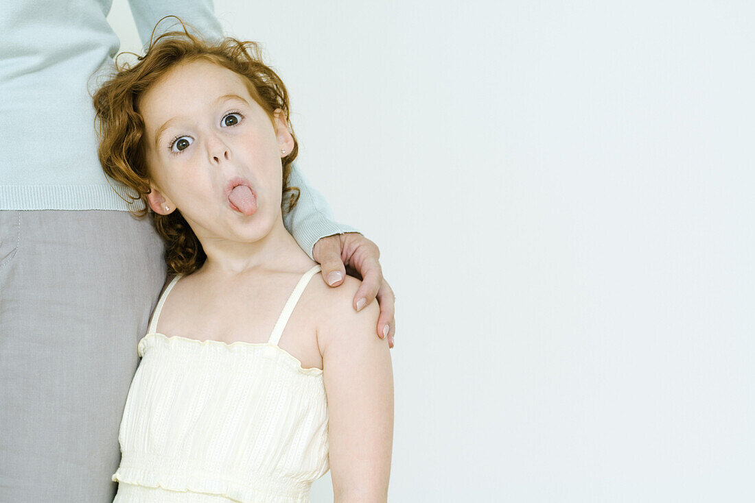 Little girl holding onto mother's leg, sticking tongue out at camera