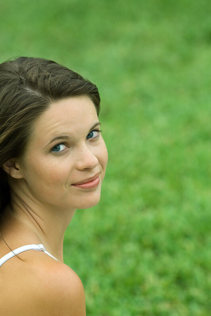 Teen girl looking over shoulder at camera, cropped, head and shoulders, grass in background