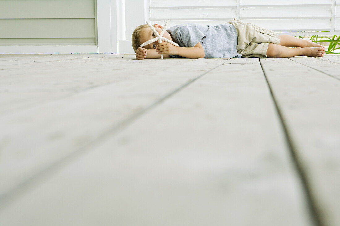 Boy lying on the ground holding starfish in front of face, looking at camera, viewed from surface level