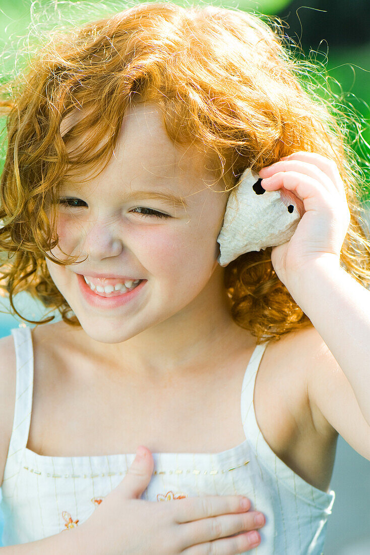 Little girl holding seashell up to ear, smiling, looking away