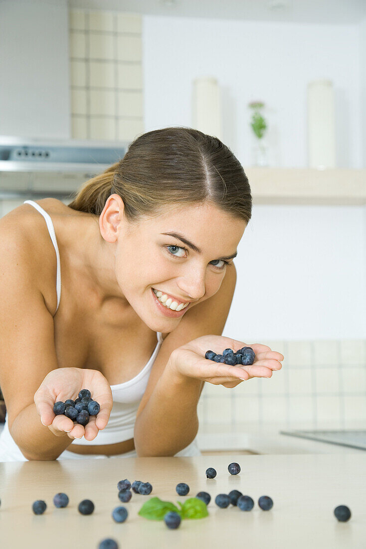 Woman in kitchen, holding handfuls of blueberries, smiling at camera