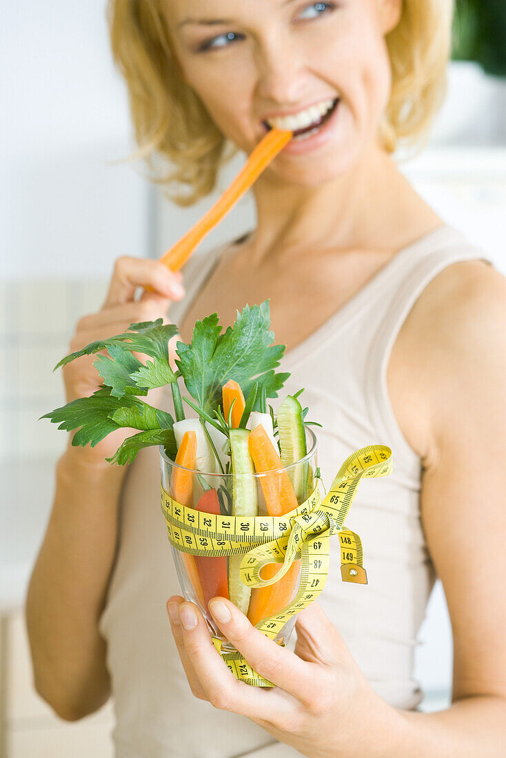 Woman biting into carrot, holding glass full of vegetables tied with measuring tape