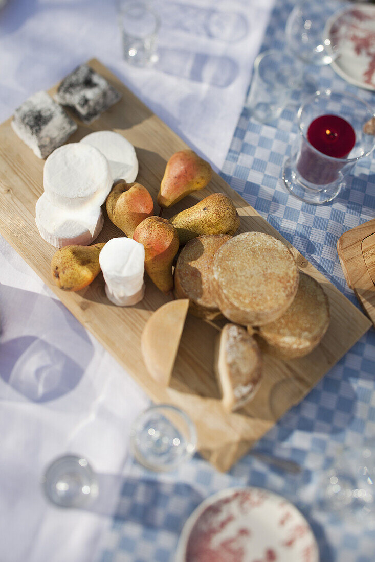 Variety of cheeses on a chopping board sitting on a table outside among glasses.