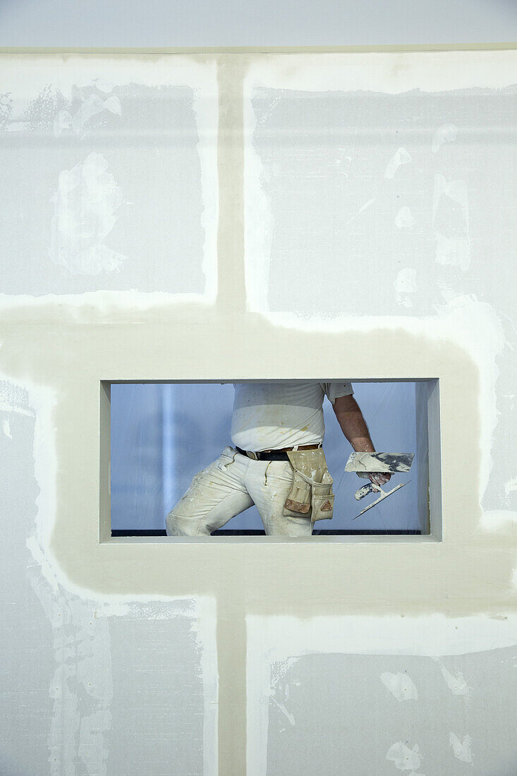 A house painter holding a trowel, seen through window hole in a newly constructed wall