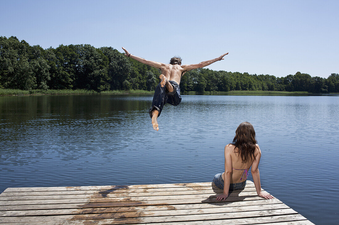 Guy dives into lake with arms outstretched as girl watches on jetty
