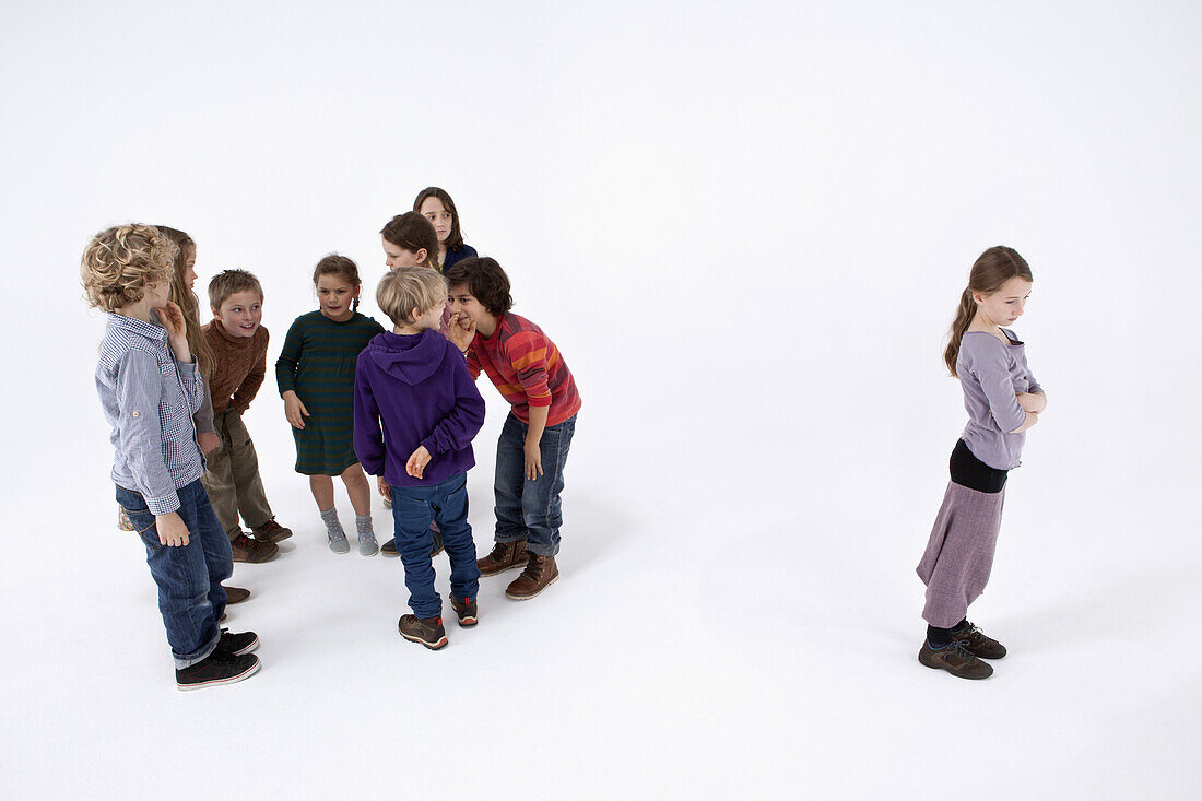 A girl standing alone, arms crossed, while a group of kids gossip about her