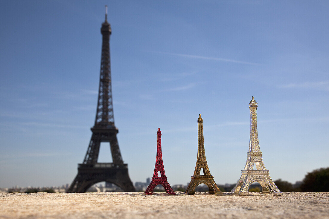 Three Eiffel Tower replica souvenirs next to the real Eiffel Tower, focus on foreground