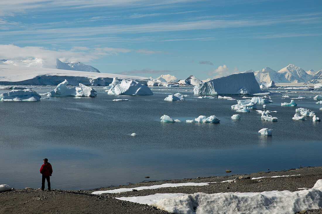 View from Rothera Station to the surrounding snowy mountains with a man standing on a hillside overlooking bay with icebergs, Rothera Station, Marguerite Bay, Antarctica