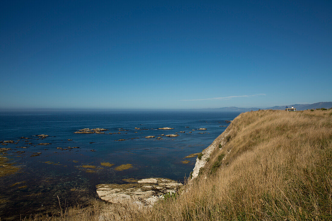 View of cliff and South Bay coastline from Peninsula Walkway with two people sitting on a bench in distance, near Kaikoura, Canterbury, South Island, New Zealand