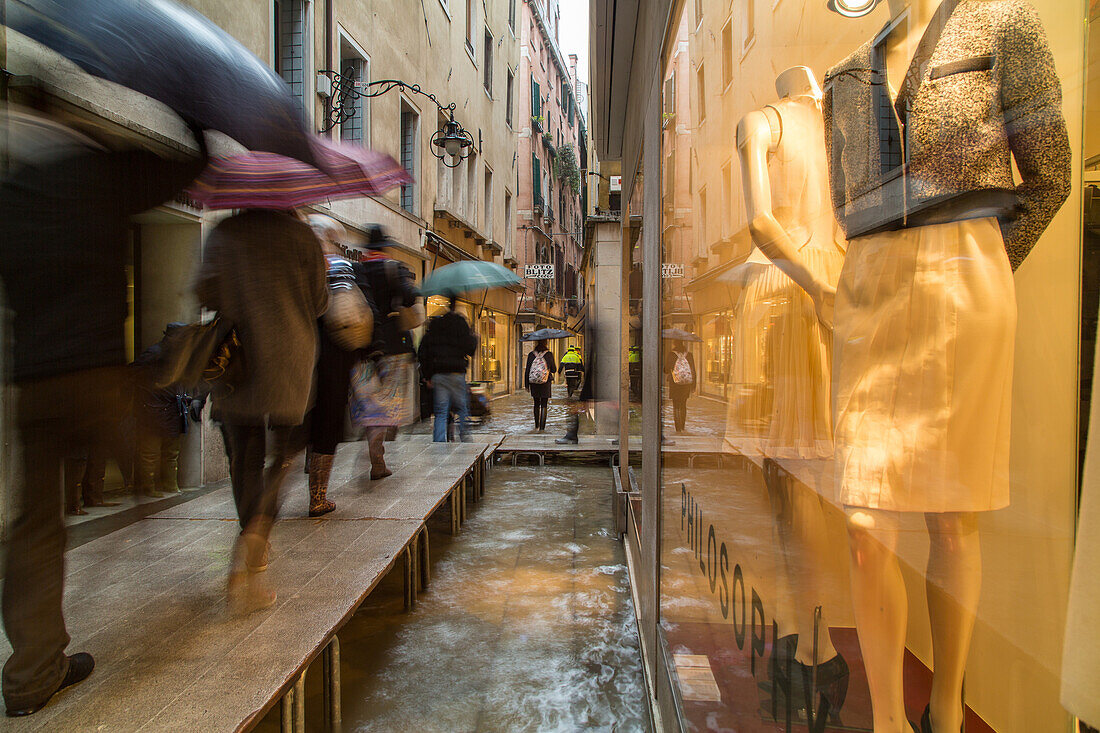 Tourists with umbrellas and plastic covered shoes, rubber boots, walk on passarelle, walkways during Acqua alta fashion shop near St Mark's Square, San Marco, passarelle, high water caused by Sirocco wind and full moon, rain in Venice, Italy