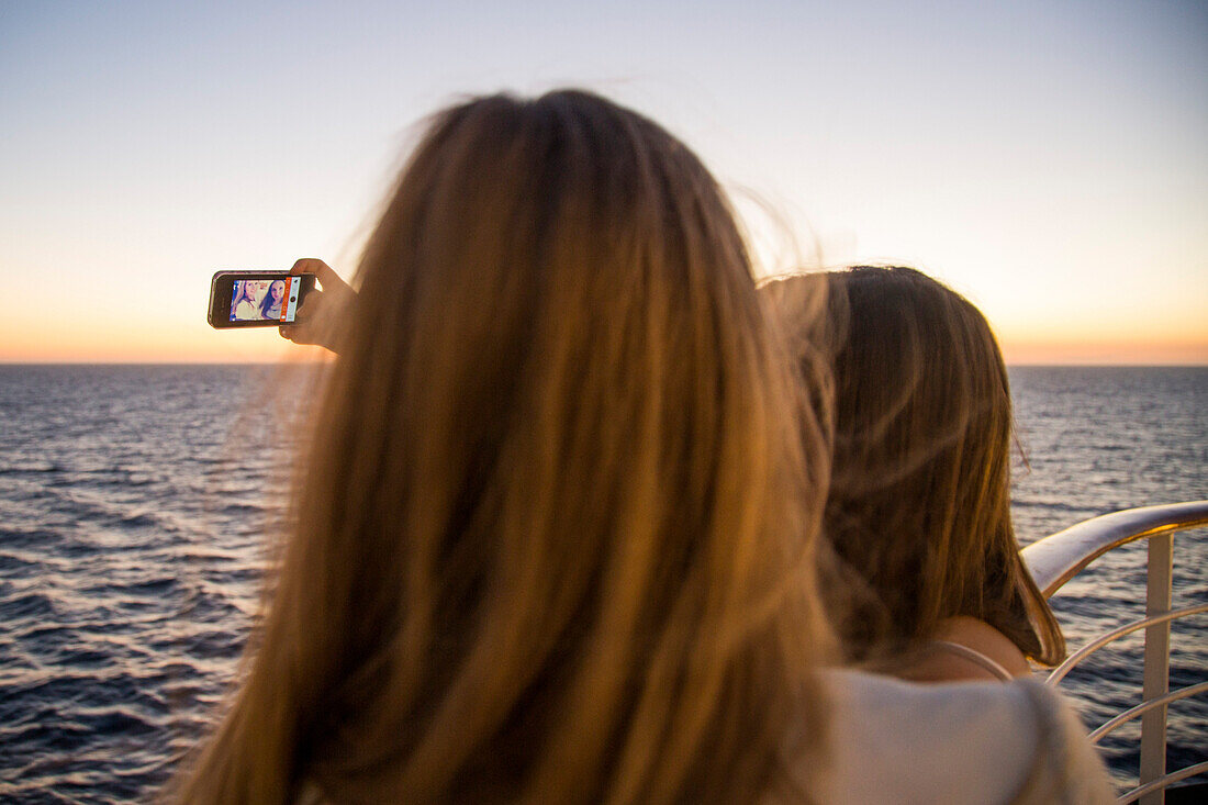 Zwei Teenager Mädchen machen ein Selfie mit ihrem Smartphone an Bord von Kreuzfahrtschiff MS Deutschland (Reederei Peter Deilmann) bei Sonnenuntergang, Atlantischer Ozean, nahe Spanien, Europa