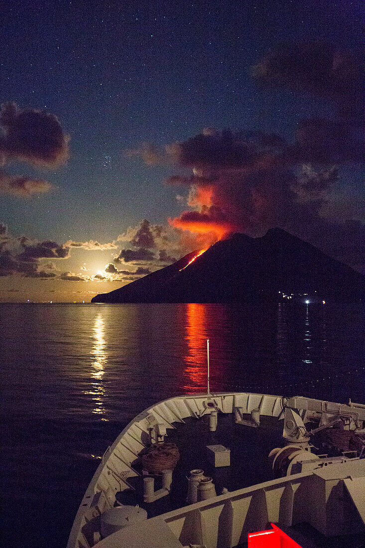 Bug von Kreuzfahrtschiff MS Deutschland (Reederei Peter Deilmann) vor aktivem Vulkan Stromboli mit Lavastrom und Mondaufgang bei Nacht, Mittelmeer, nahe Lipari, Äolische Inseln, nahe Sizilien, Italien, Europa