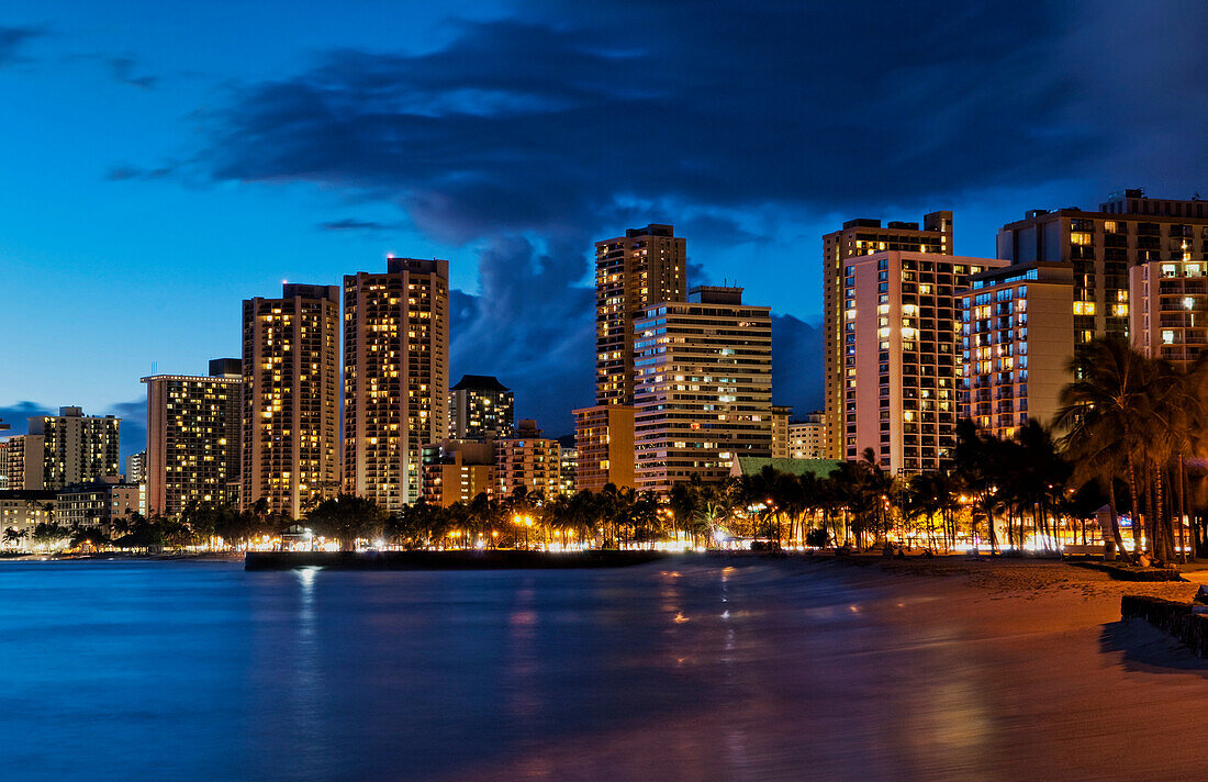 Hawaii, Oahu, Waikiki, Waikiki Beach At Night With Veiw Of City Lights And Buildings.