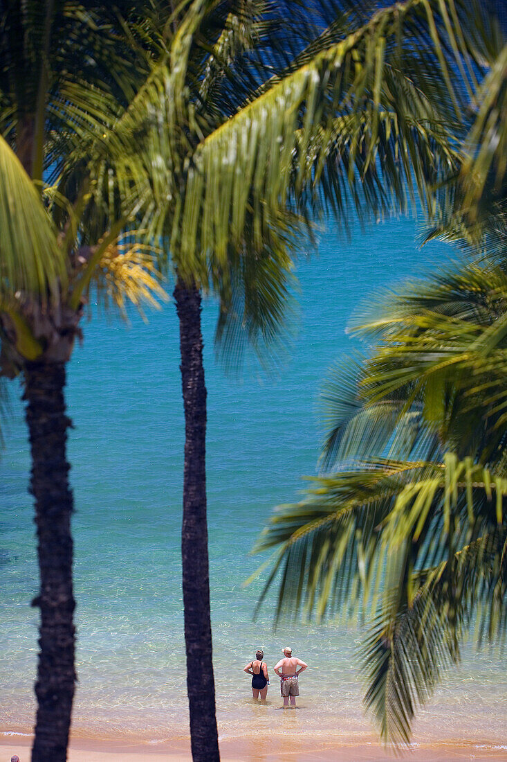 Hawaii, Maui, Wailea, Mokapu Beach, Senior Couple Stands Knee Deep In Water Looking Out At Ocean, Palm Trees Surround.