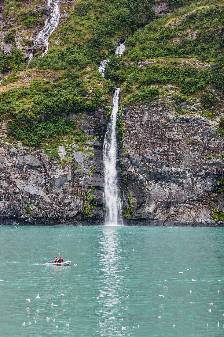 Waterfall spilling over a cliff into Passage canal with a small boat in the foreground, Prince William Sound, Whittier Southcentral Alaska, USA.
