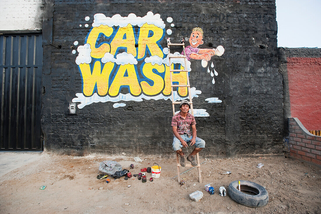 'A man sits on a ladder in front of his painted car wash sign on a brick wall; Chimbote, Peru'