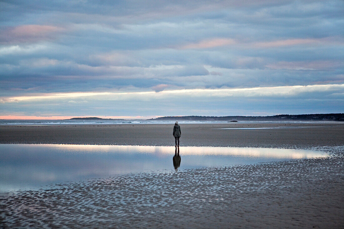 A woman stands reflected in a tidal pool at a beach in Maine.