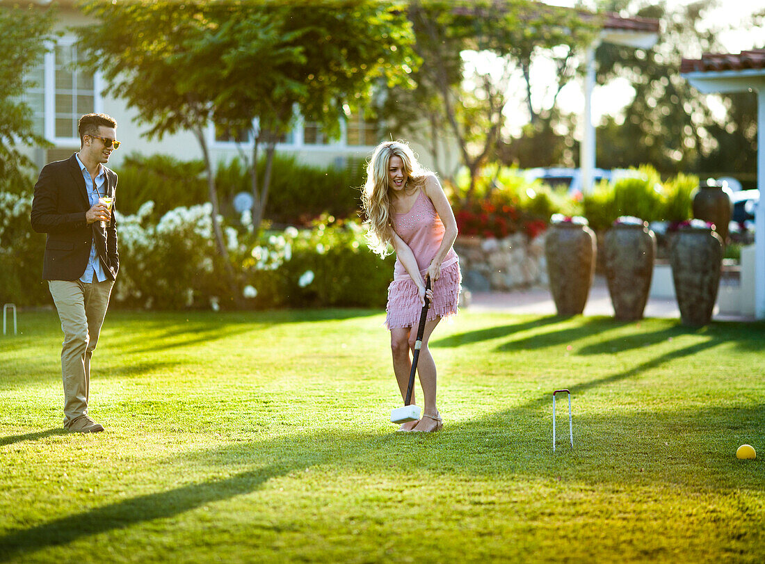 A beautiful lady hits a croquet ball through a wicket, her male companion stands on her side enjoys it with a drink.