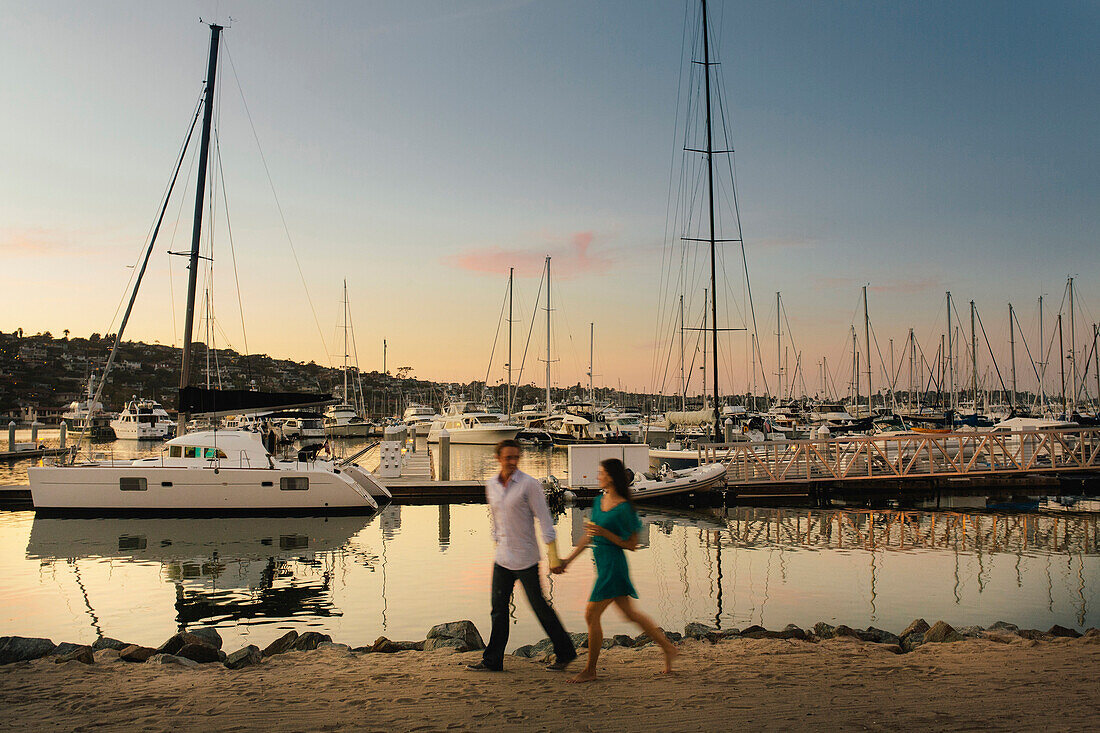 A young girl with a drink in her left hand strolls by the marina along her male partner hand in hand.