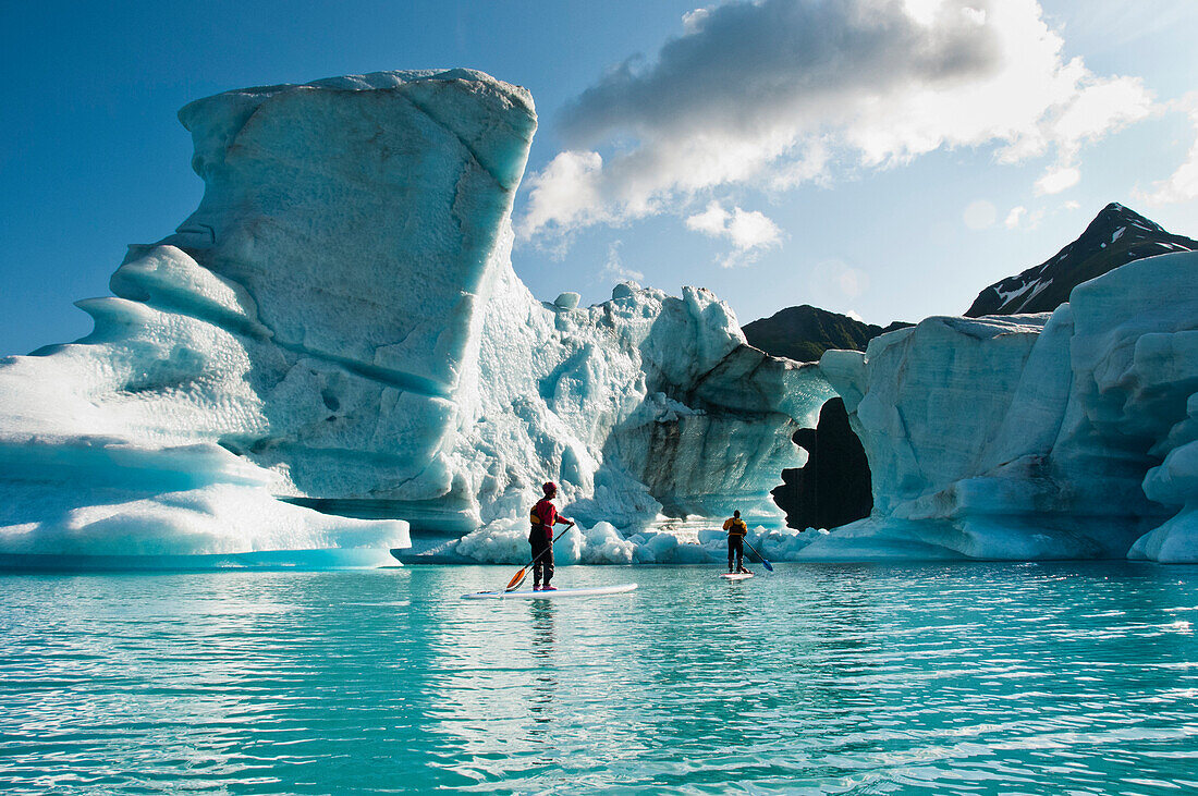 Two adults on stand up paddle board (SUP) observe hole melted in iceberg on Bear Lake in Kenai Fjords National Park, Alaska.