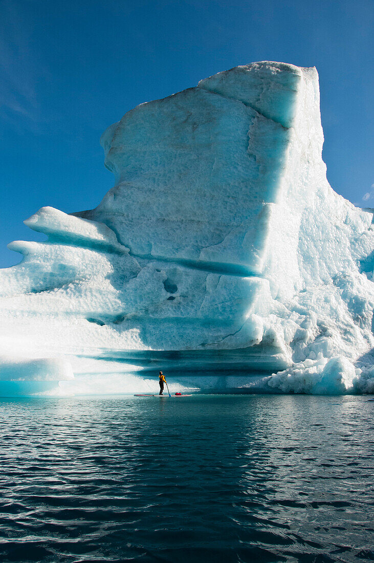 A man stand up paddle boards (SUP) in front of a towering iceberg on Bear Lake in Kenai Fjords National Park, Alaska.