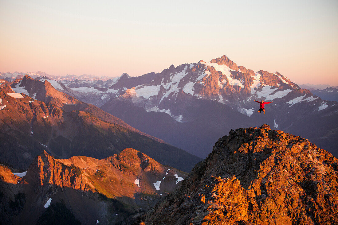 A hiker jumps into the air from a rocky ridge in the North Cascade Mountains with Mount Shuksan in the background.
