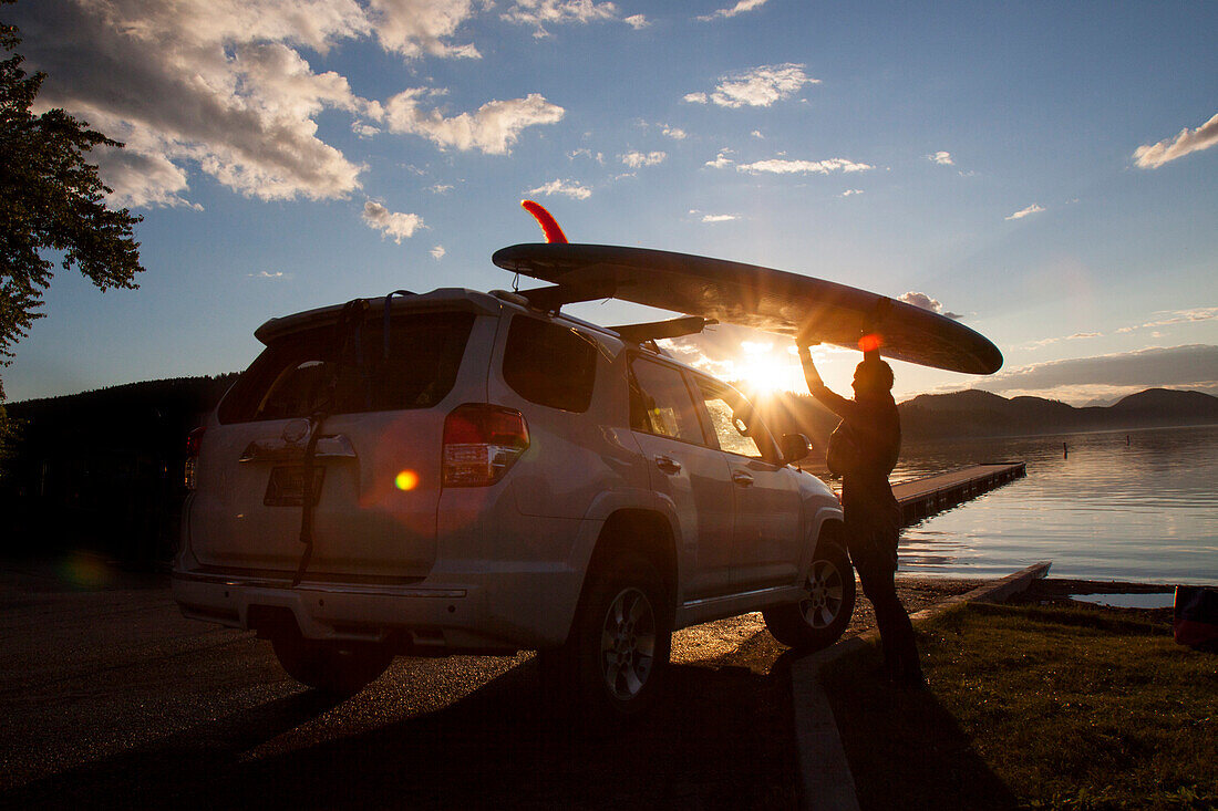 A fit female places her stand up paddle board (SUP) on a car at sunset on Whitefish Lake in Whitefish, Montana.