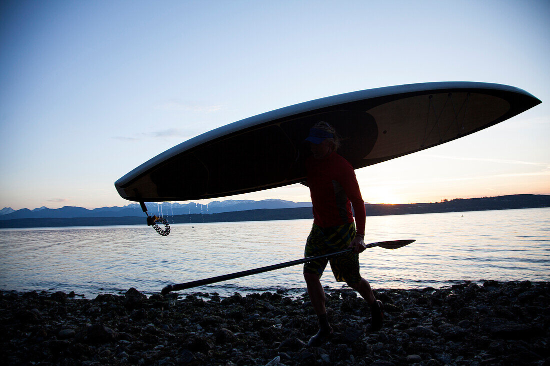 A fit male carries his stand up paddle board (SUP) along the Hood Canal in the Puget Sound with the Olympic Mountains behind him near Poulsbo, Washington at sunset.