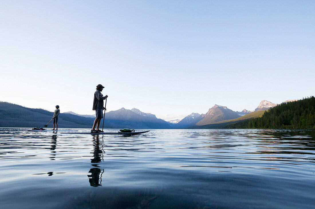 A man and woman stand up paddle boards (SUP) on Lake McDonald at sunset in Glacier National Park near West Glacier, Montana.
