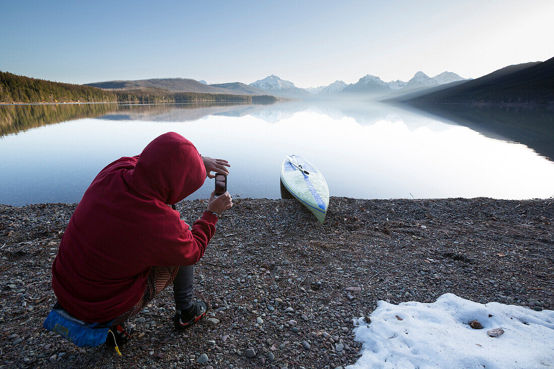 A man takes a photograph with his smart phone while stand up paddle boarding (SUP) on a calm Lake McDonald at sunrise in Glacier National Park near West Glacier, Montana.