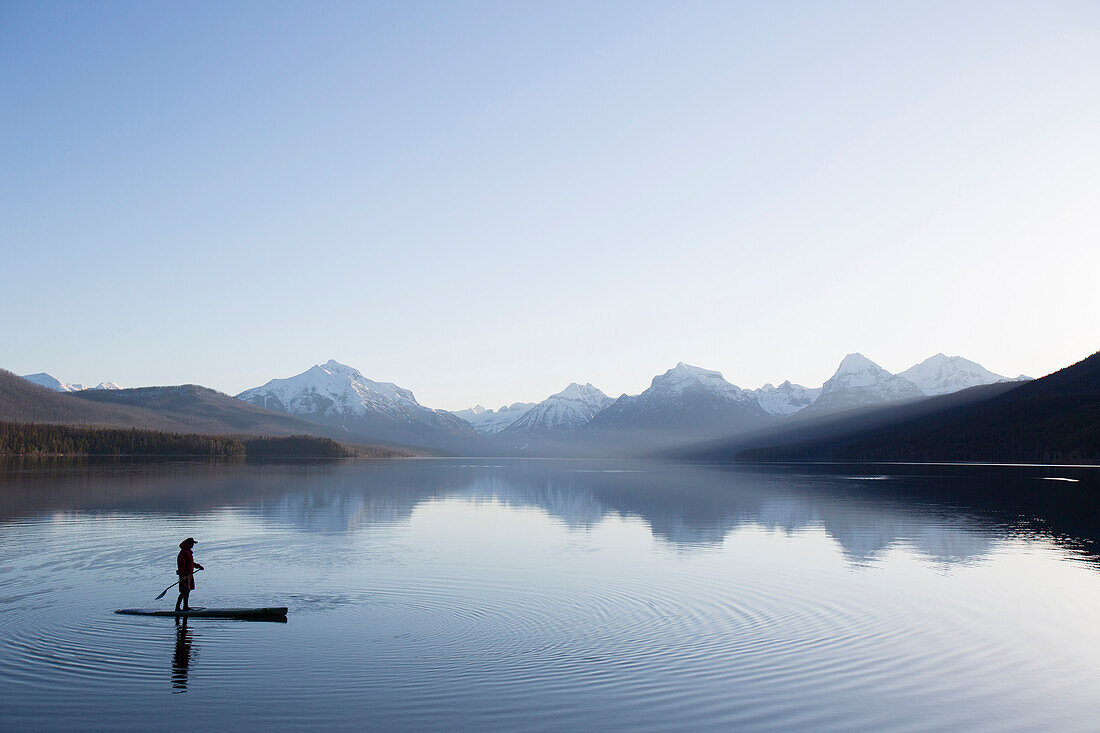 A man stand up paddle boards (SUP) on a calm Lake McDonald at sunrise in Glacier National Park near West Glacier, Montana.