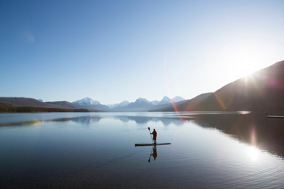 A man stand up paddle boards (SUP) on a calm Lake McDonald at sunrise in Glacier National Park near West Glacier, Montana.