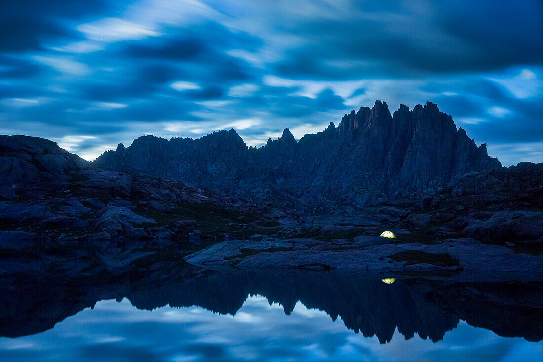 An illuminated tent reflected in a lake in front of Jagged peak deep in the Weminuche wilderness near Silverton Colorado.