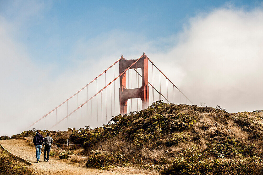 The Golden Gate surrounded by fog, partially visible over a hill where two guys are walking.