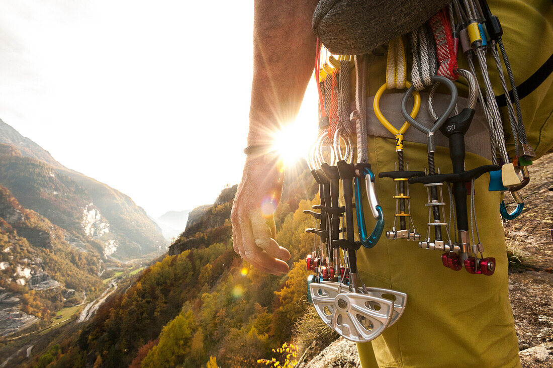 Various climbing gear including friends and nuts hanging on a climber's harness, whyle the climber stands at the top of a cliff with a view of Formazza Valley, backlit by the sun. Cadarese, Ossola, Italy.