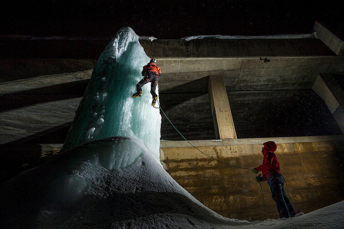 Two boys climbing an icefall near a tunnel on Simplon Pass road by night, lit with headlamps and strobes.
