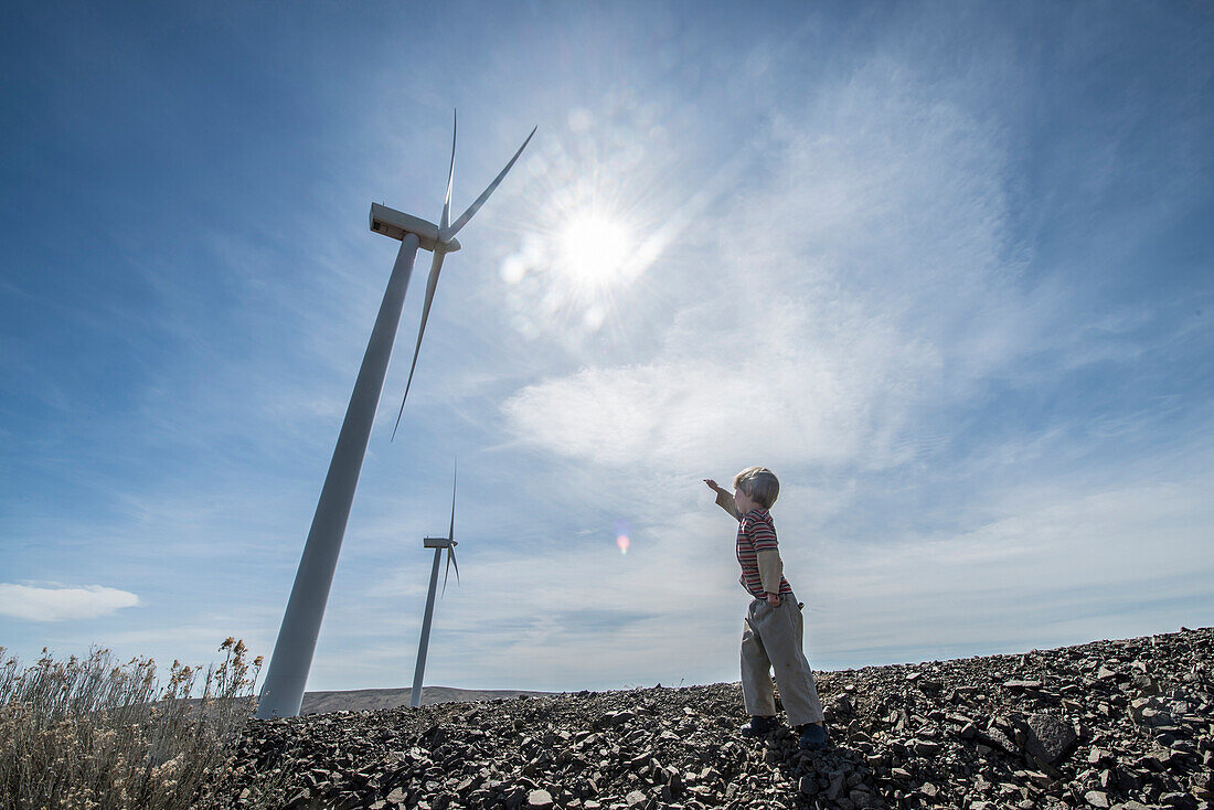 A young boy points up to a wind turbine with the sun in the background.