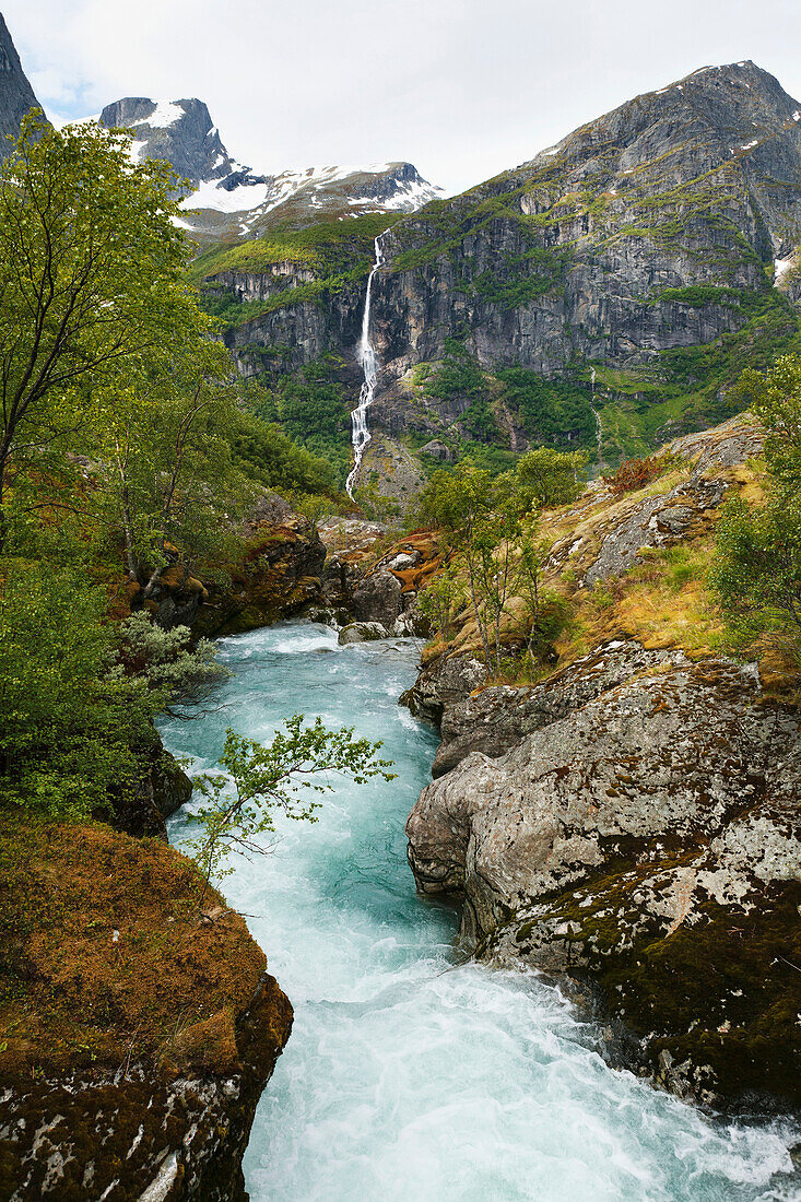 'Water rushing in a river in a valley between the mountains with a waterfall on a steep cliff in the distance; Olden, Norway'