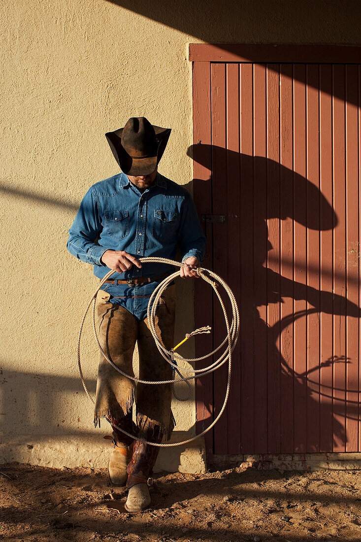 Agriculture - Cowboy with a lasso rope standing against a barn in evening light / Childress, Texas, USA.