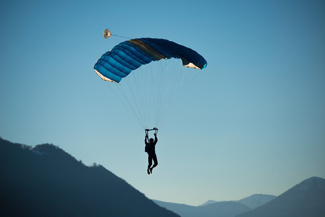 'A skydiver floats with a parachute against a blue sky and the swiss alps; Locarno, Ticino, Switzerland'