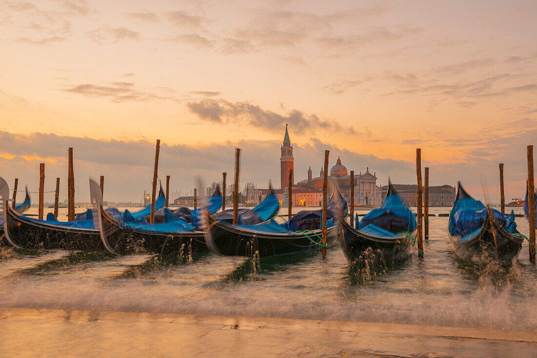 'Gondolas mooring at the shore of the Grand Canal with waves splashing water at sunset; Venice, Veneto, Italy'