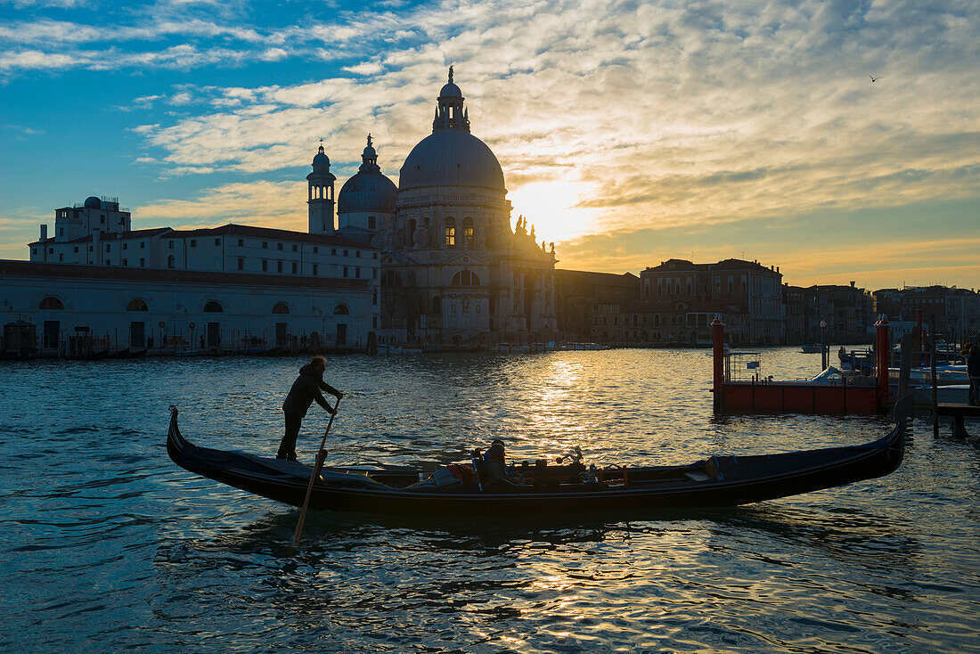 'Silhouette of a gondolier rowing his gondola with Salute church along the shoreline in the distance at sunset; Venice, Veneto, Italy'
