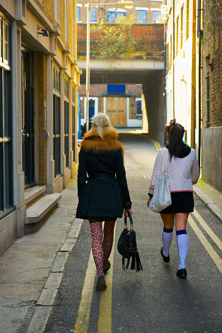 'Two young women walking down a narrow street between buildings, Brick Lane; London, England'