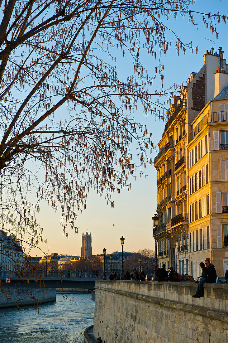'A couple sit together on a canal wall with sunset sunlight cast on the buildings; Paris, France'