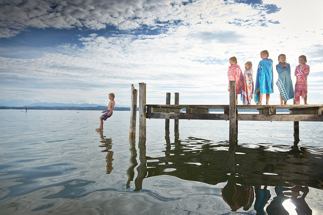 Children wrapped in towels on a jetty, boy jumping into the water, lake Starnberg, Upper Bavaria, Bavaria, Germany